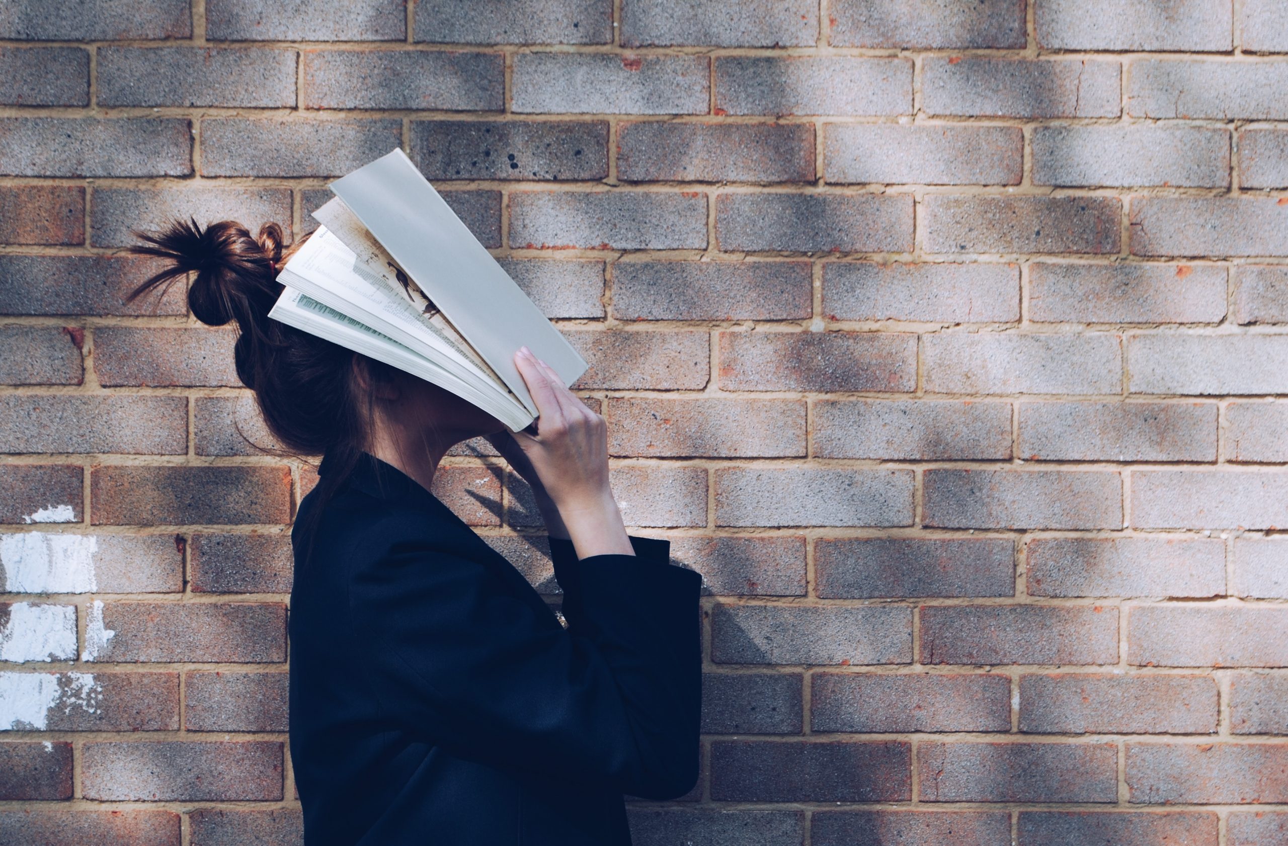 Girl standing by brick wall with open book covering her face