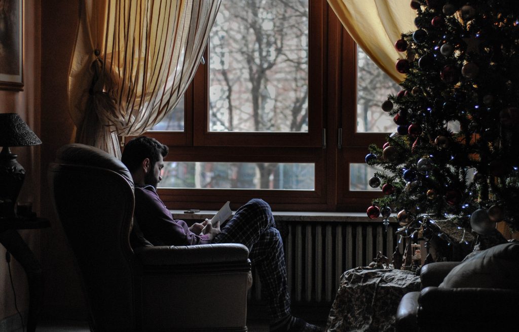 Man sitting by christmas tree and window reading book in pyjamas